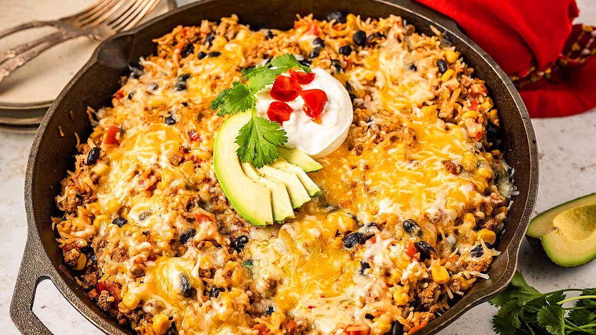 A skillet filled with cheesy taco casserole topped with sour cream, avocado slices, cilantro, and diced red peppers. Plates and a red napkin are in the background.