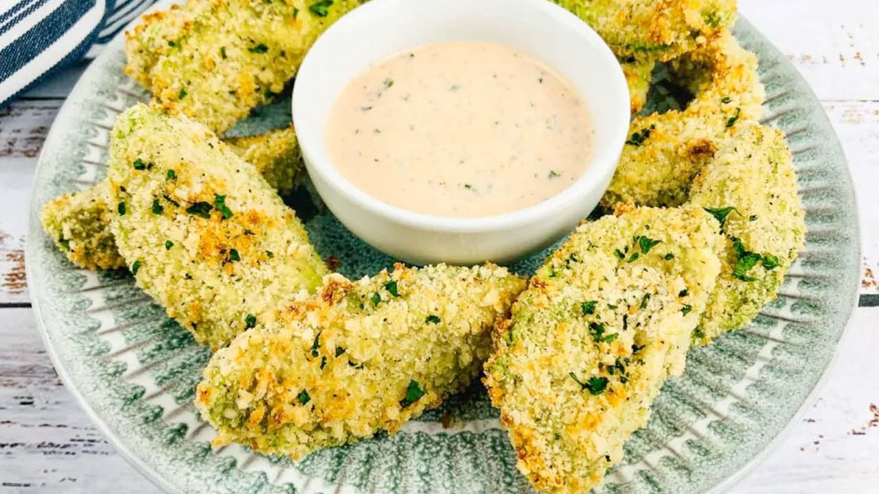 A plate of breaded avocado fries garnished with herbs is arranged around a bowl of creamy dipping sauce.