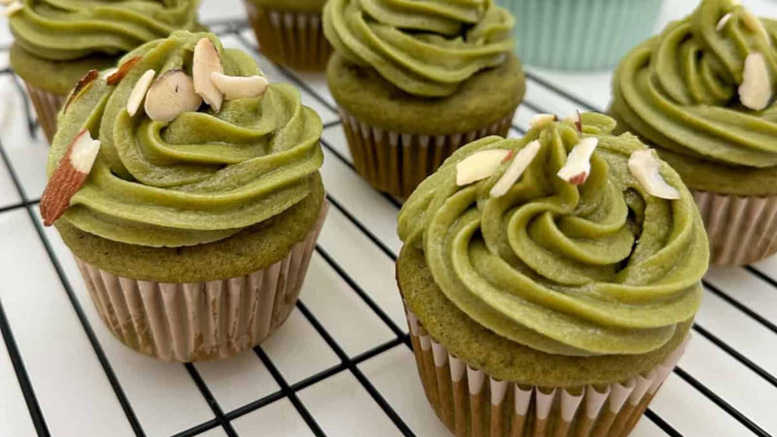 Green frosted cupcakes with almond slices on top, arranged on a cooling rack.
