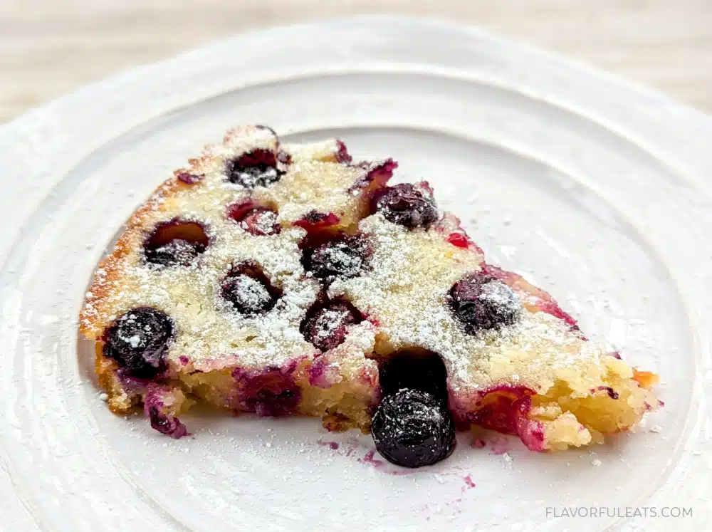 A slice of blueberry clafoutis dusted with powdered sugar sits on a white plate.