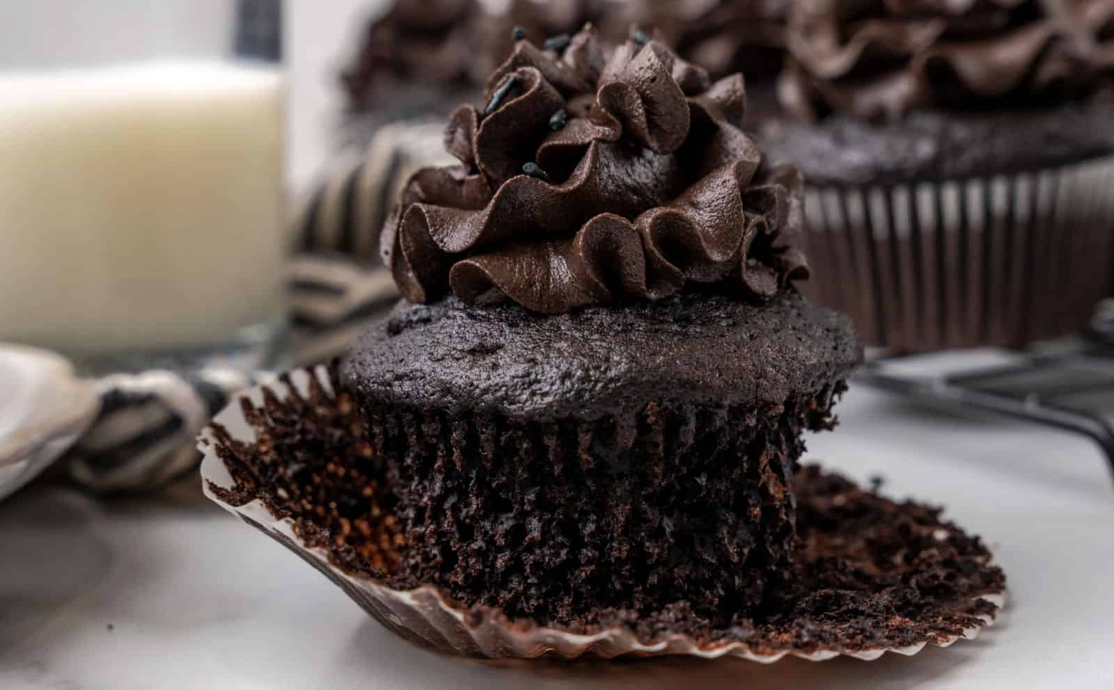 A partially unwrapped chocolate cupcake with dark chocolate frosting sits on a marble surface, with a glass of milk and more cupcakes in the background.