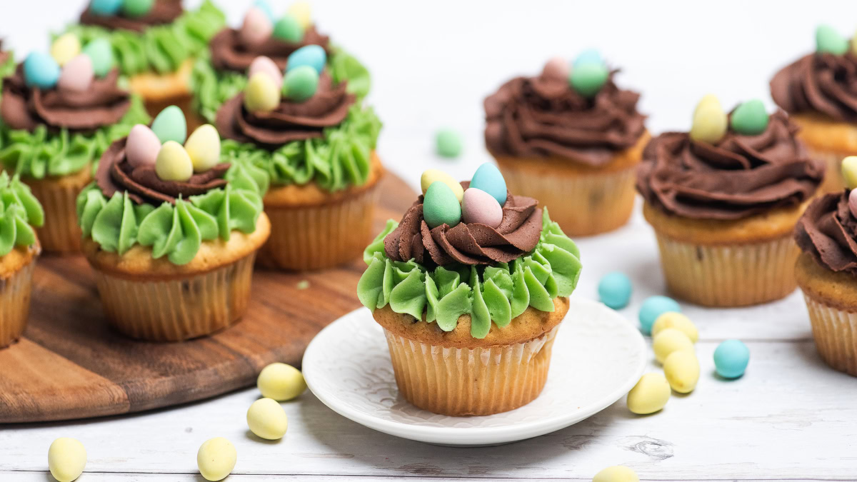 Vanilla cupcakes with green and chocolate frosting are topped with small pastel candy eggs, arranged on a wooden board and a white plate, with extra candies scattered around.