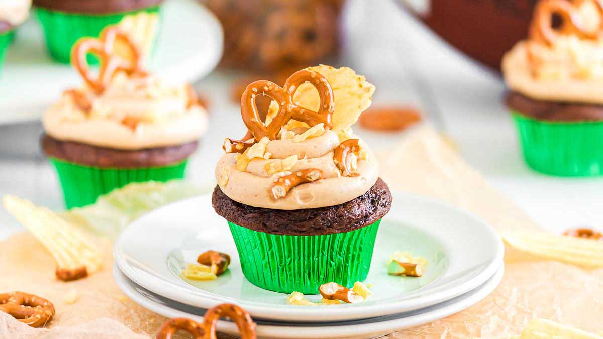 A chocolate cupcake in a green wrapper topped with light brown frosting, pretzels, and crushed pretzel pieces sits on a white plate with more cupcakes in the background.