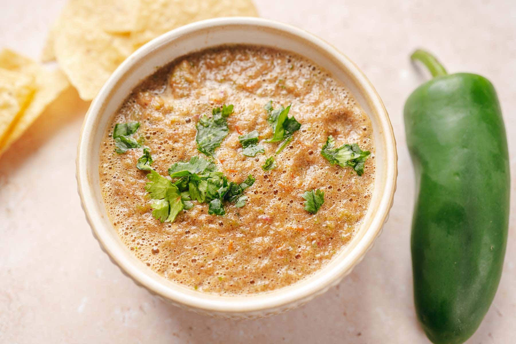 A bowl of blended salsa garnished with chopped cilantro, next to a whole jalapeรฑo pepper and tortilla chips.
