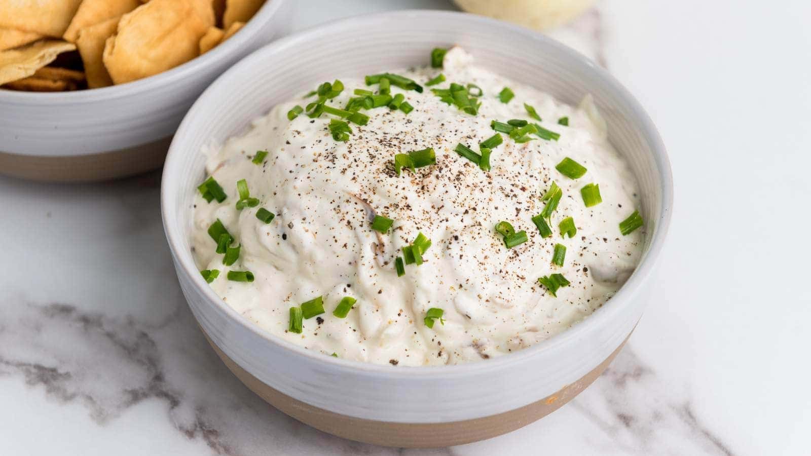 A bowl of creamy dip topped with chopped chives and cracked black pepper, with a bowl of crackers nearby on a marble surface.