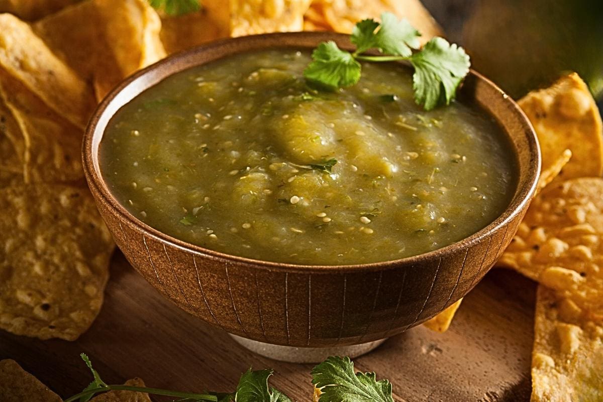 A brown bowl filled with green tomatillo salsa garnished with cilantro, surrounded by yellow tortilla chips on a wooden surface.
