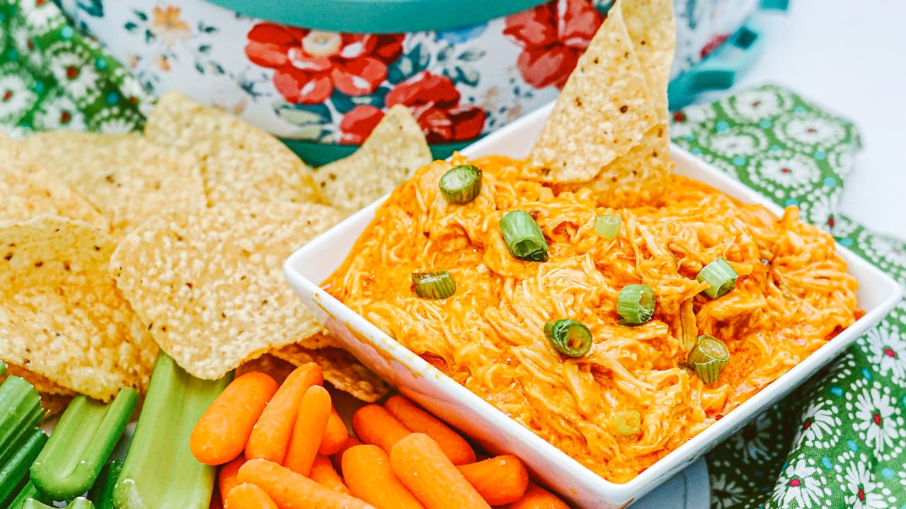 A square bowl of buffalo chicken dip topped with chopped green onions, surrounded by tortilla chips, celery sticks, and baby carrots.