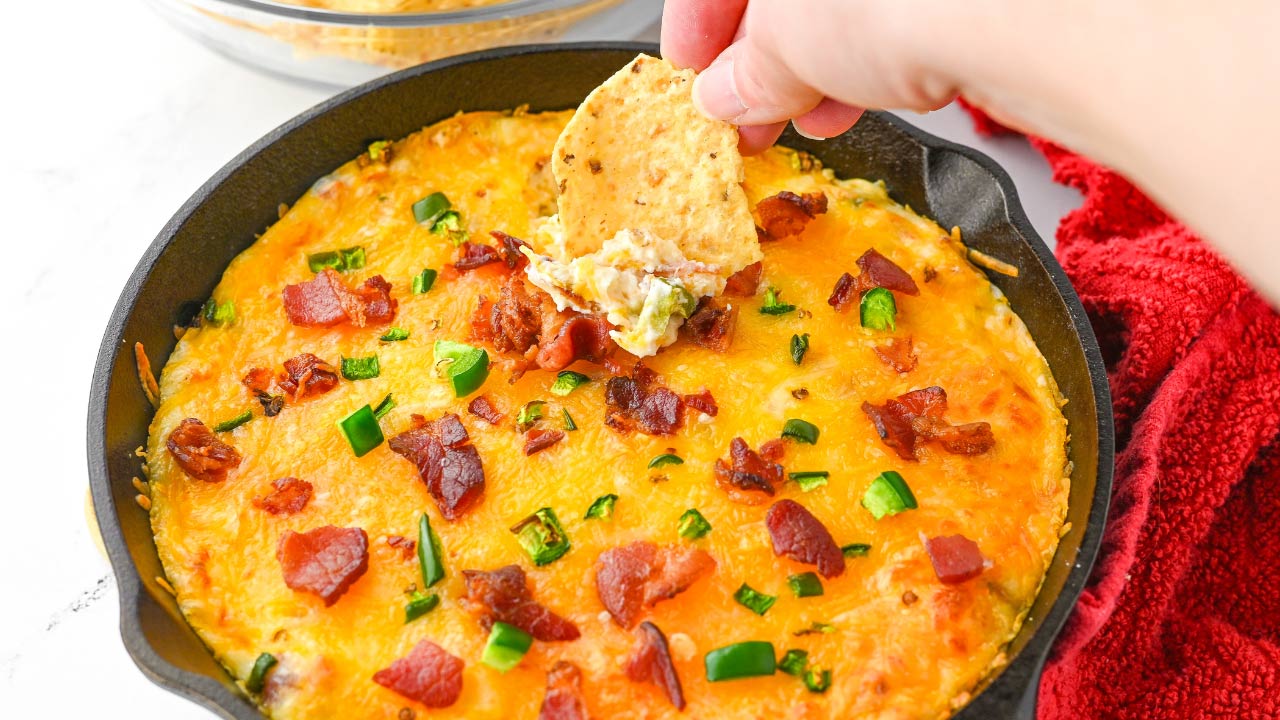 A hand dips a tortilla chip into a cast iron skillet filled with cheesy dip topped with chopped bacon and green peppers.