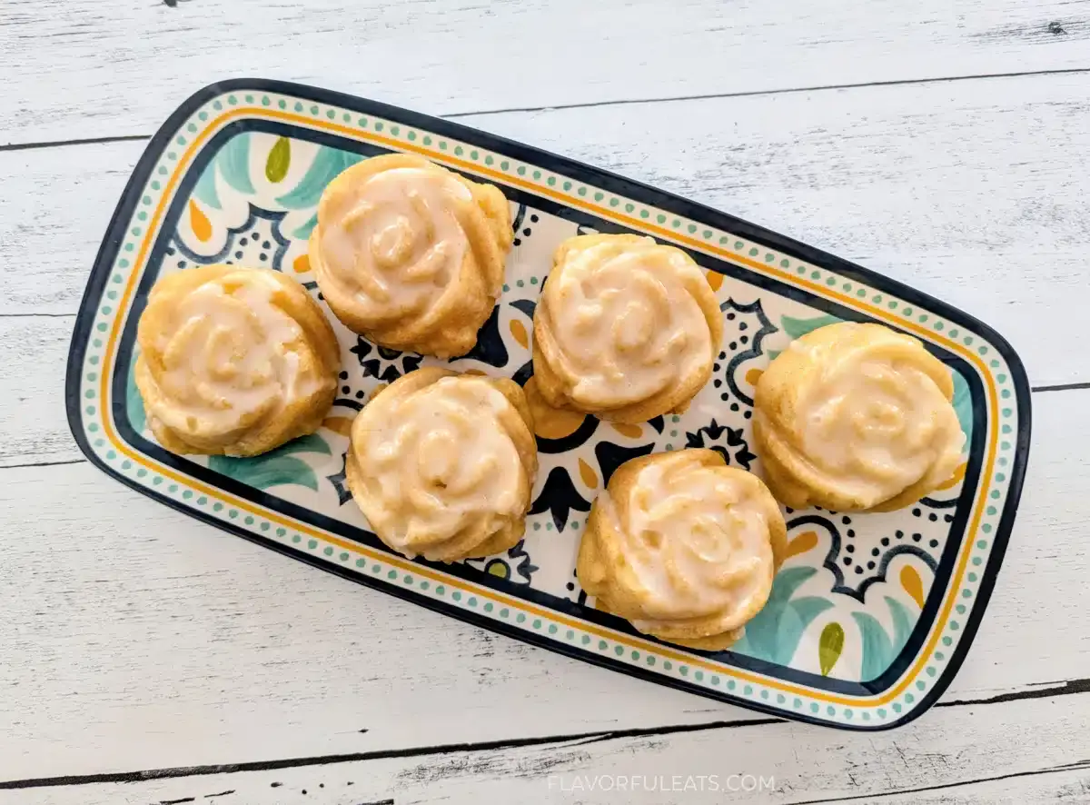 A rectangular patterned plate holds six round pastries with light icing, arranged in two rows on a white wooden surface.