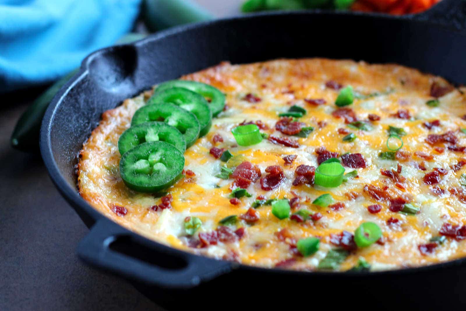 A baked frittata in a cast iron skillet, topped with sliced jalapeรฑos, chopped green onions, melted cheese, and bacon bits.