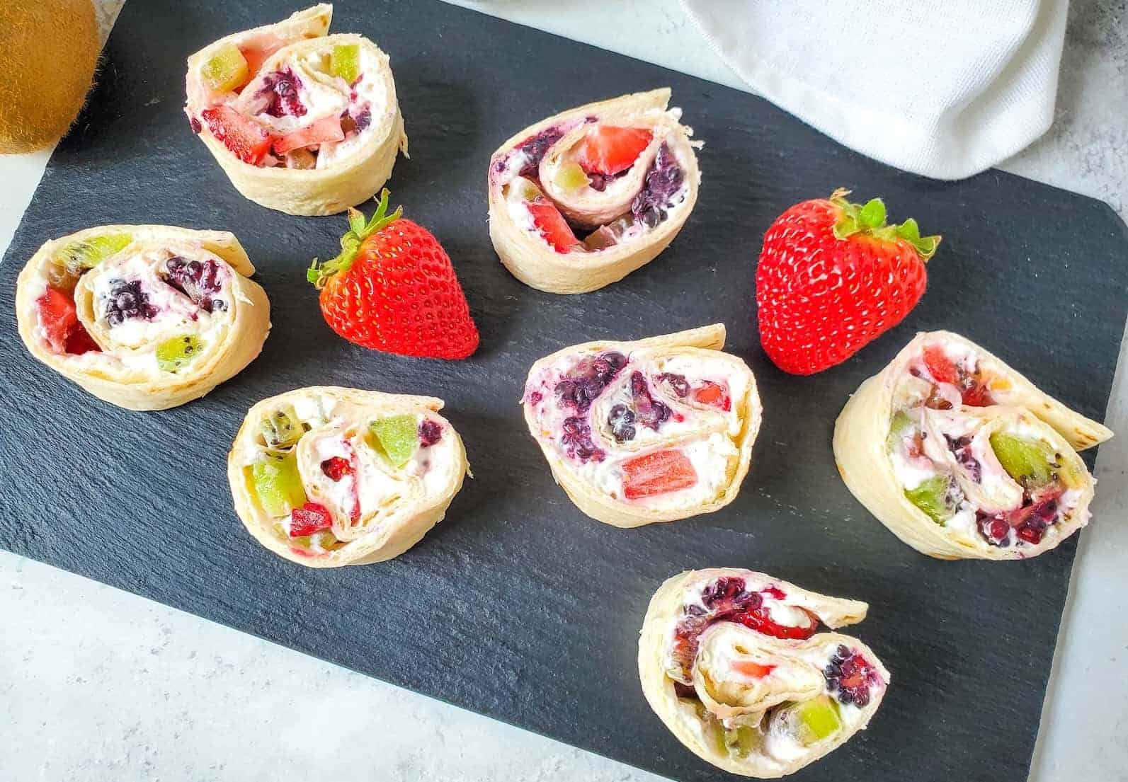 Sliced fruit and cream cheese tortilla pinwheels arranged on a black slate board with two whole strawberries.