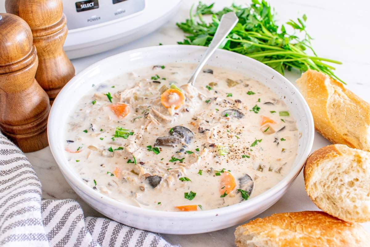 A bowl of creamy soup with mushrooms, carrots, herbs, and shredded meat, served with a spoon, fresh bread, and garnished with pepper and parsley.