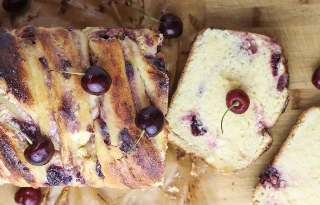 A loaf of braided bread with cherries on top and visible cherry pieces inside, shown on a wooden surface with slices cut.