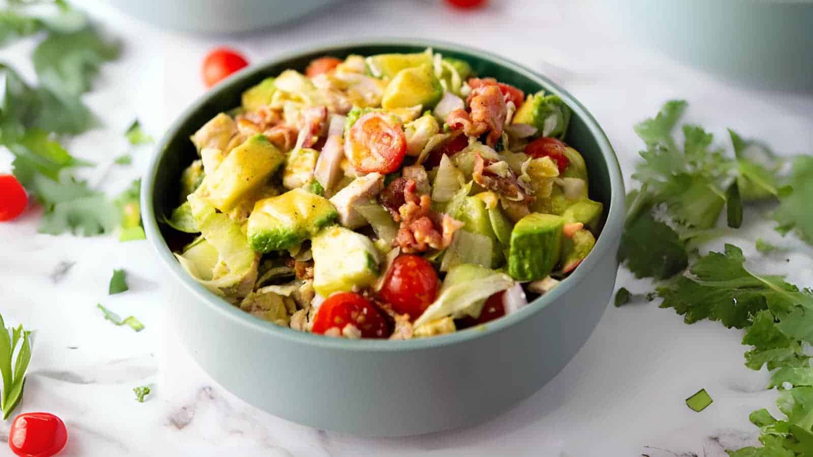 A bowl of chopped salad with lettuce, cherry tomatoes, avocado, bacon, and dressing, placed on a marble surface with herbs scattered around.