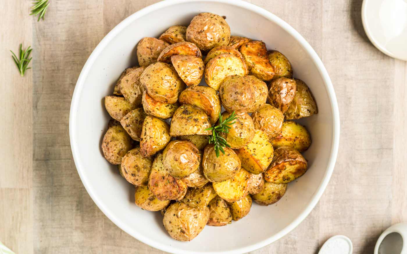 A white bowl filled with roasted baby potatoes seasoned with herbs, garnished with a sprig of rosemary, placed on a light-colored surface.