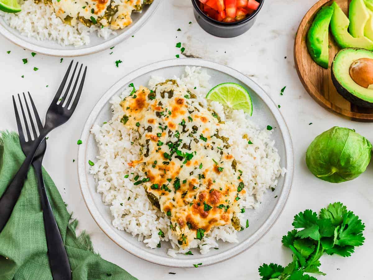 A plate of white rice topped with a cheesy green chicken casserole, garnished with cilantro and a lime wedge, with sides of fresh avocado, chopped tomatoes, and cilantro.