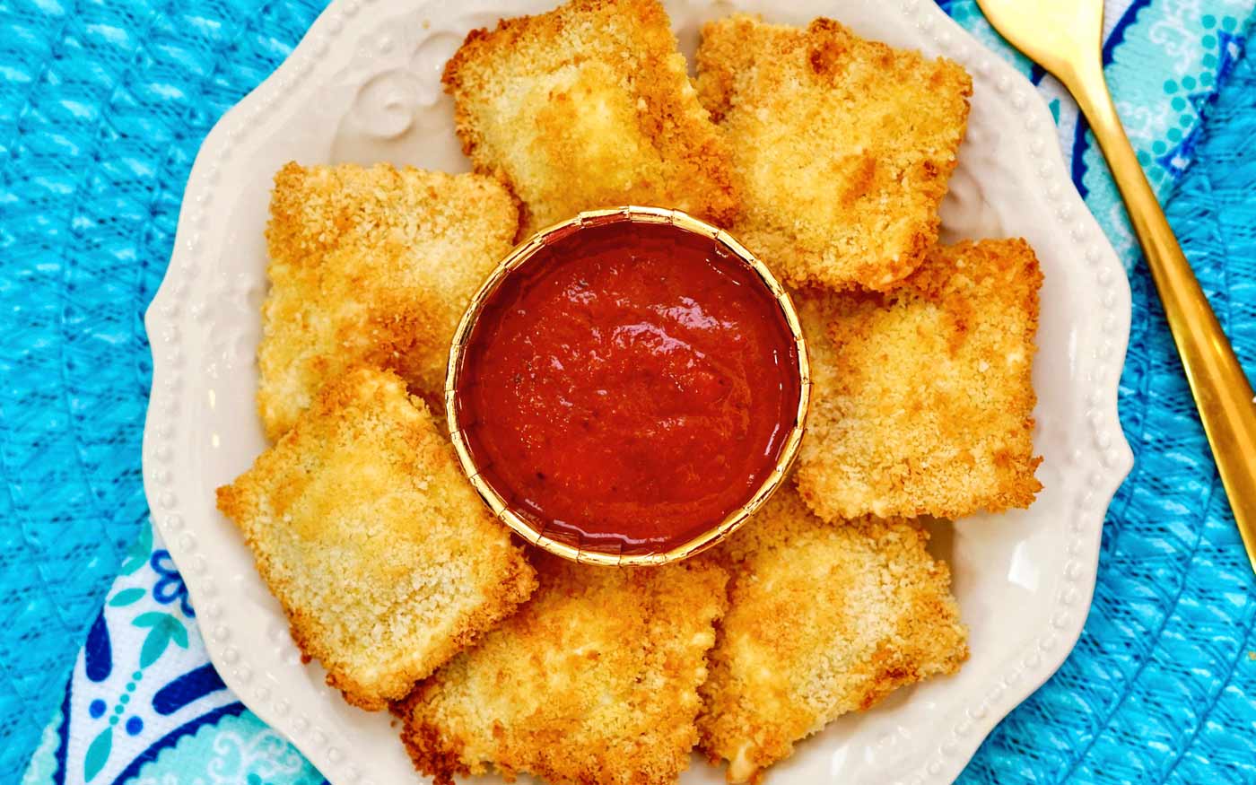 White plate with eight breaded toasted ravioli arranged around a small bowl of marinara sauce, set on a blue patterned surface with a gold fork and knife nearby.