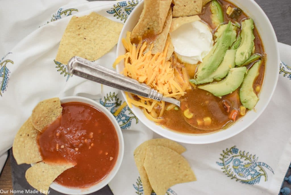 A bowl of soup with tortilla chips, shredded cheese, avocado slices, and sour cream, next to a small bowl of salsa and extra chips on a patterned cloth.