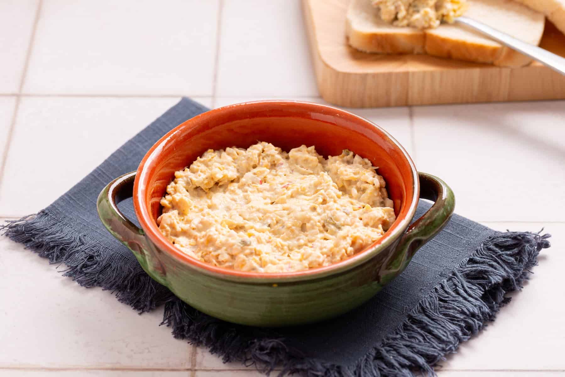 A green ceramic bowl filled with a creamy, chunky dip sits on a blue napkin. In the background, bread with some of the dip spread on it is visible.