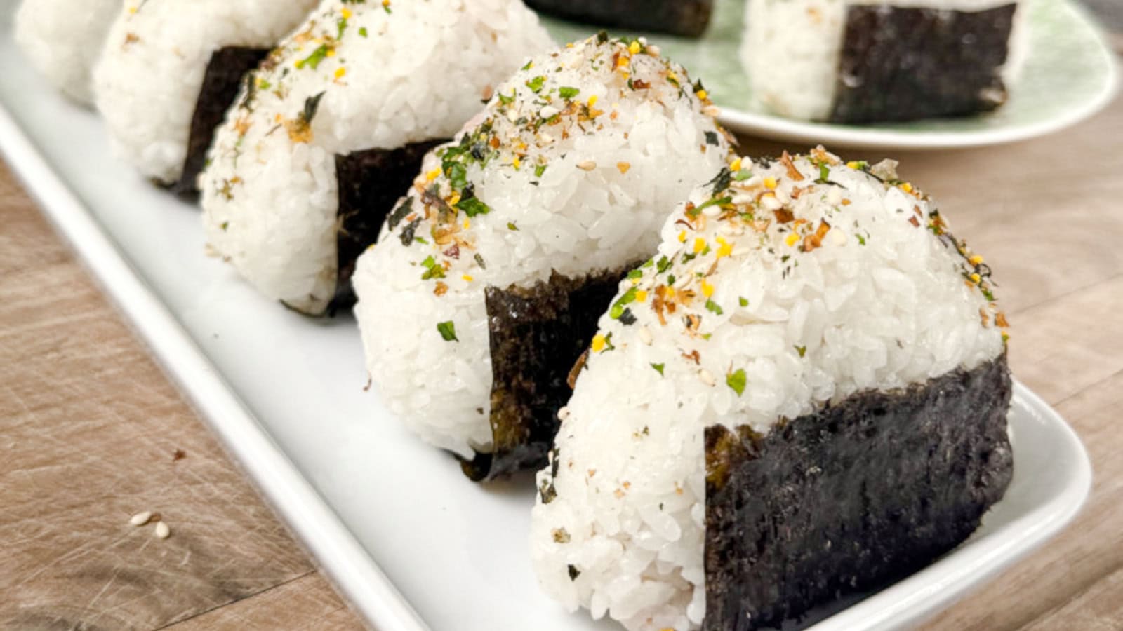 Several rice balls wrapped in seaweed and sprinkled with seasoning are arranged on a white rectangular plate on a wooden surface.