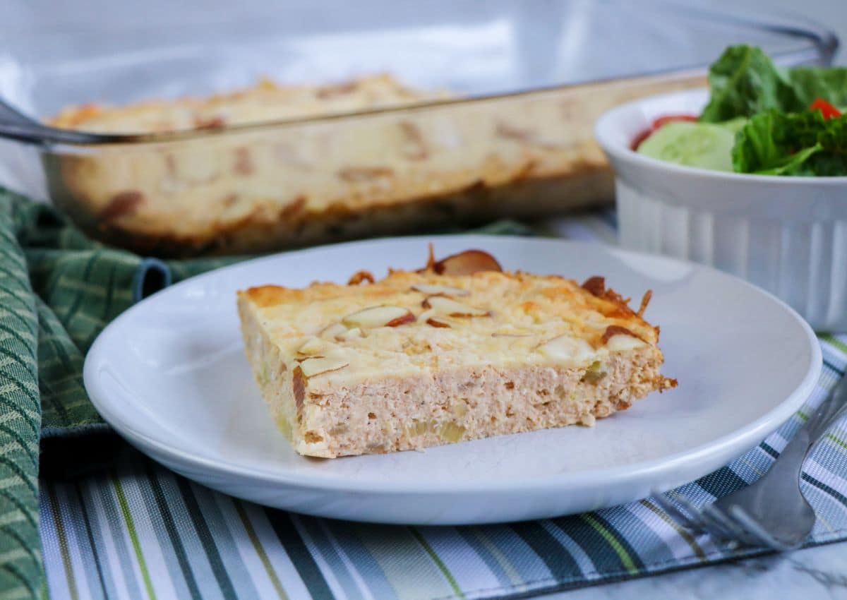 A slice of baked casserole on a white plate, with a glass baking dish and a small bowl of salad in the background.