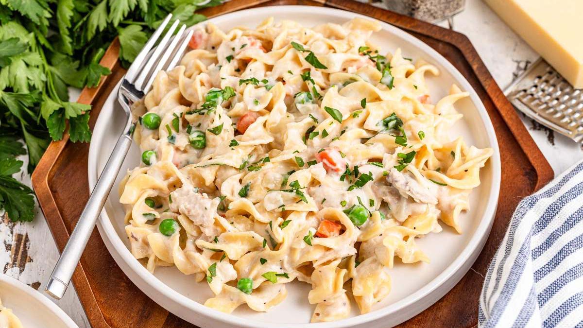 A plate of creamy pasta with peas, carrots, and shredded chicken, garnished with chopped herbs, sits on a table beside a fork and fresh parsley.