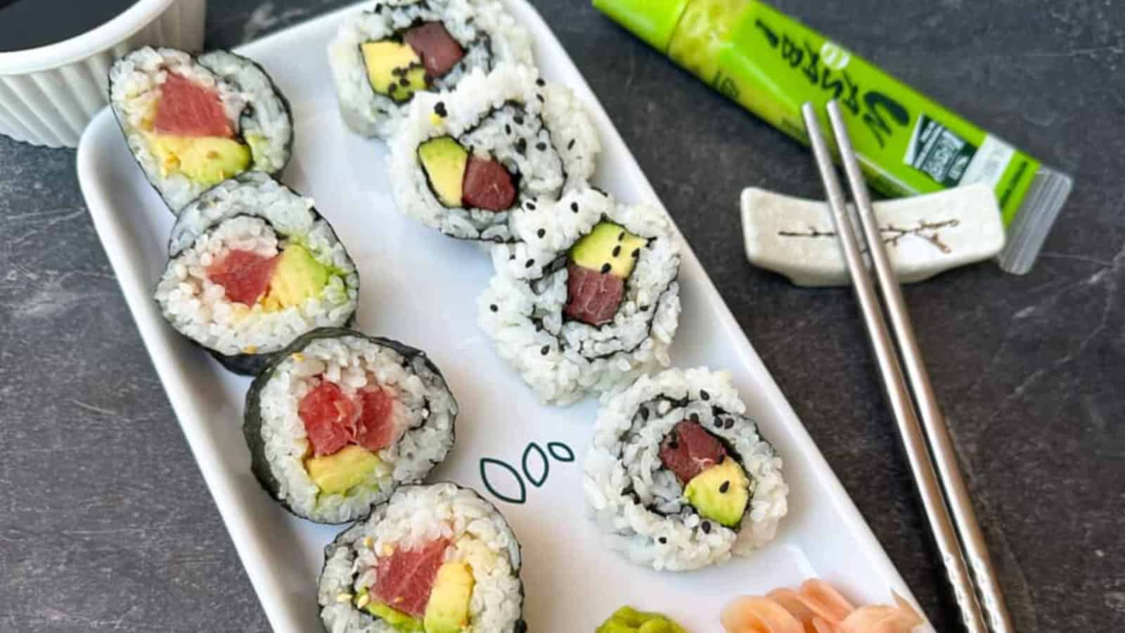 A rectangular plate with sushi rolls filled with tuna and avocado, accompanied by pickled ginger, wasabi, soy sauce, chopsticks, and a green wasabi tube on a dark surface.