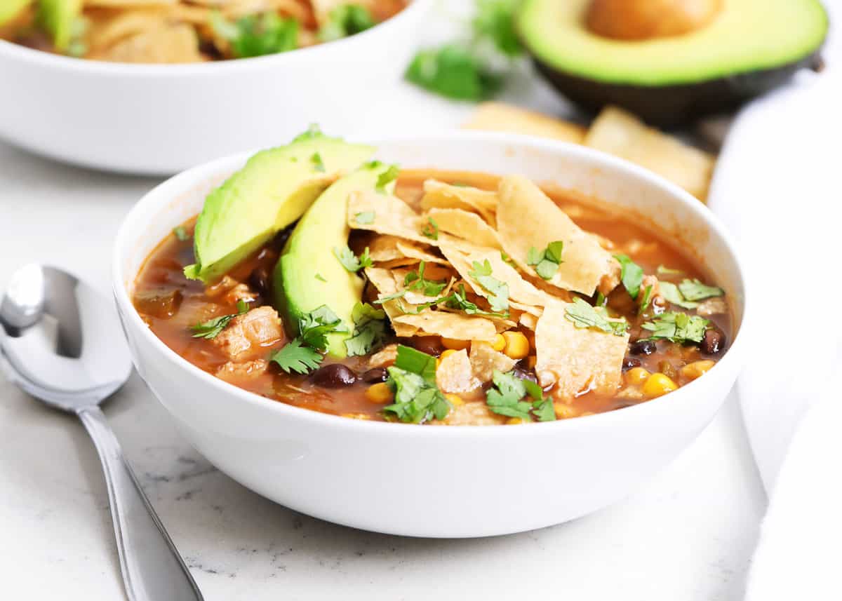 A bowl of tortilla soup topped with avocado slices, tortilla strips, and cilantro sits next to a spoon and a halved avocado.