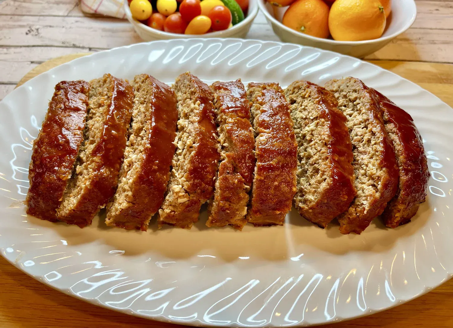 Sliced meatloaf arranged on a white oval platter, with a bowl of assorted fruits in the background.