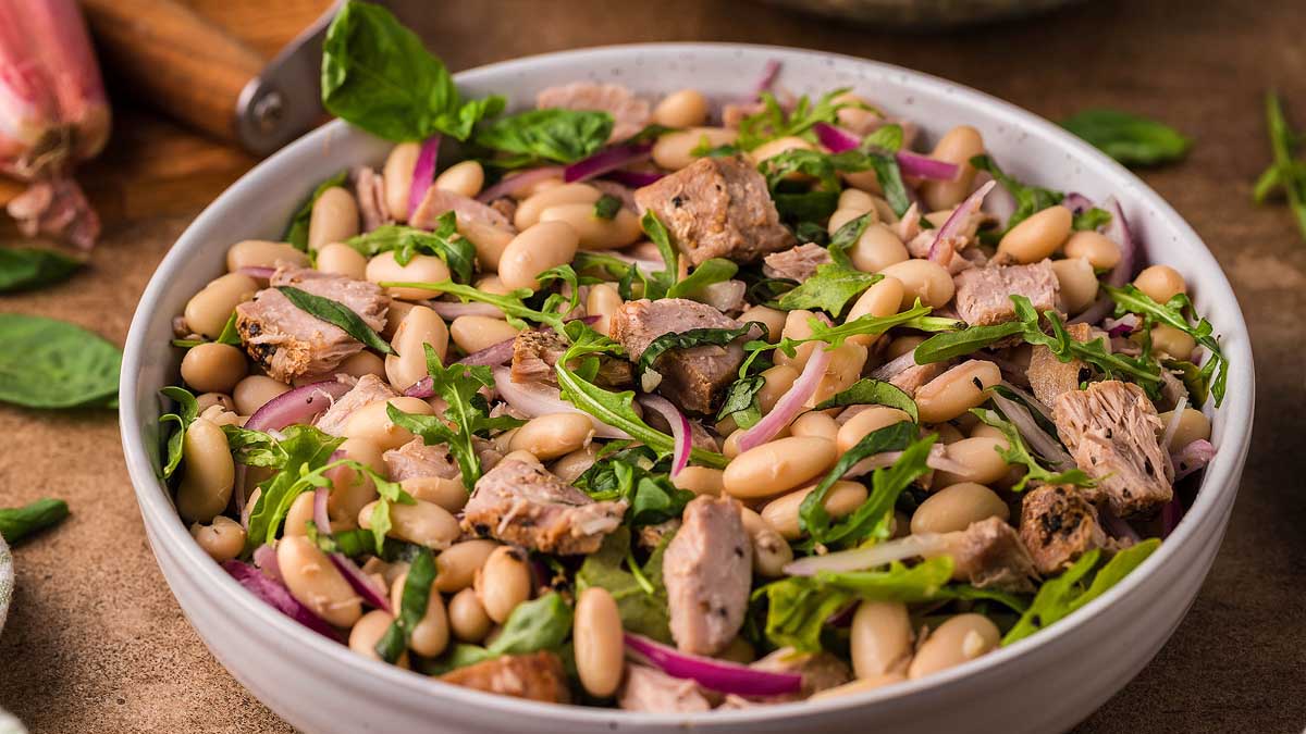 A bowl of salad containing white beans, sliced red onions, grilled meat pieces, and fresh greens.