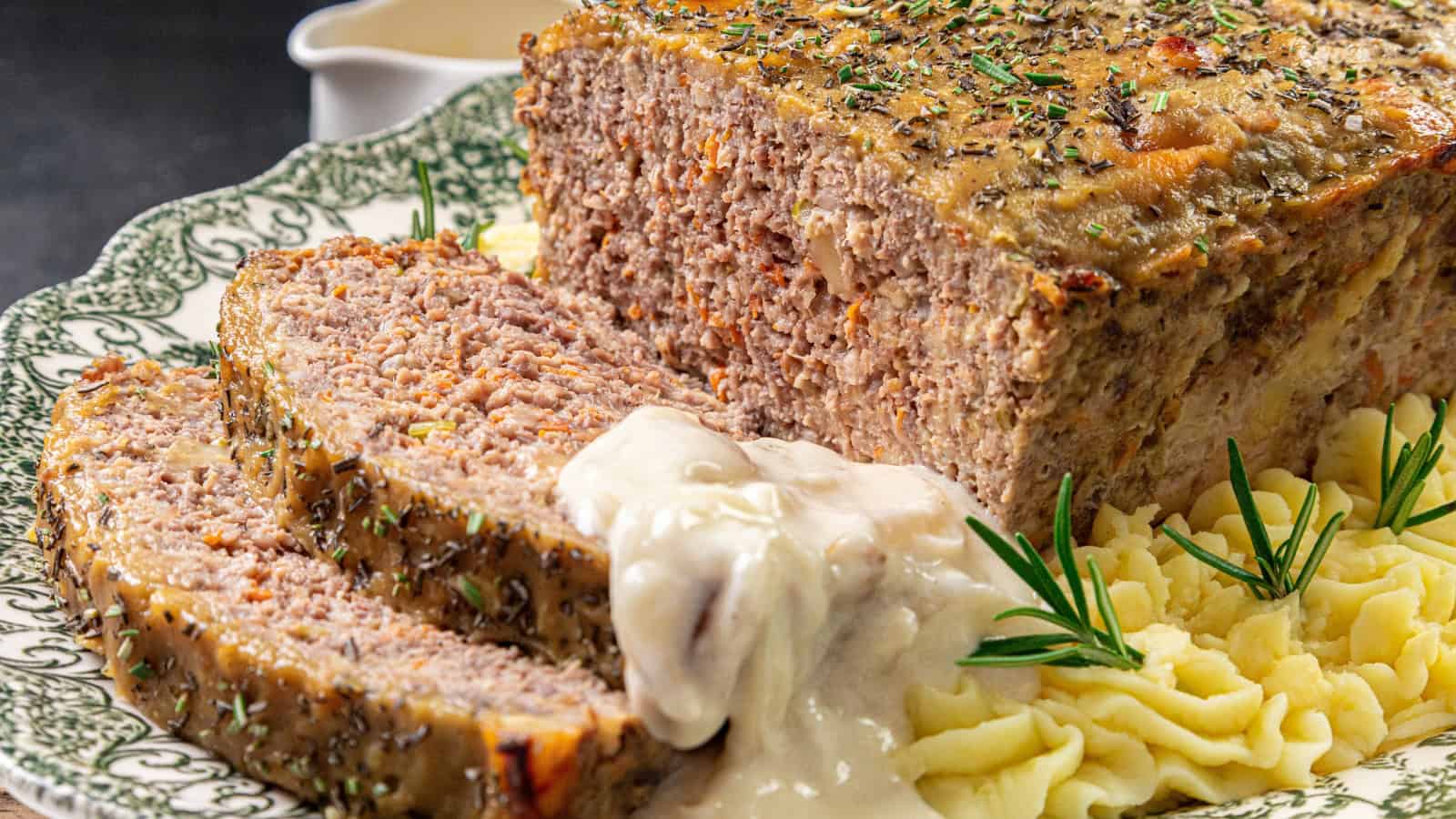 A sliced meatloaf with gravy is served on a decorative plate next to mashed potatoes, garnished with rosemary and herbs.