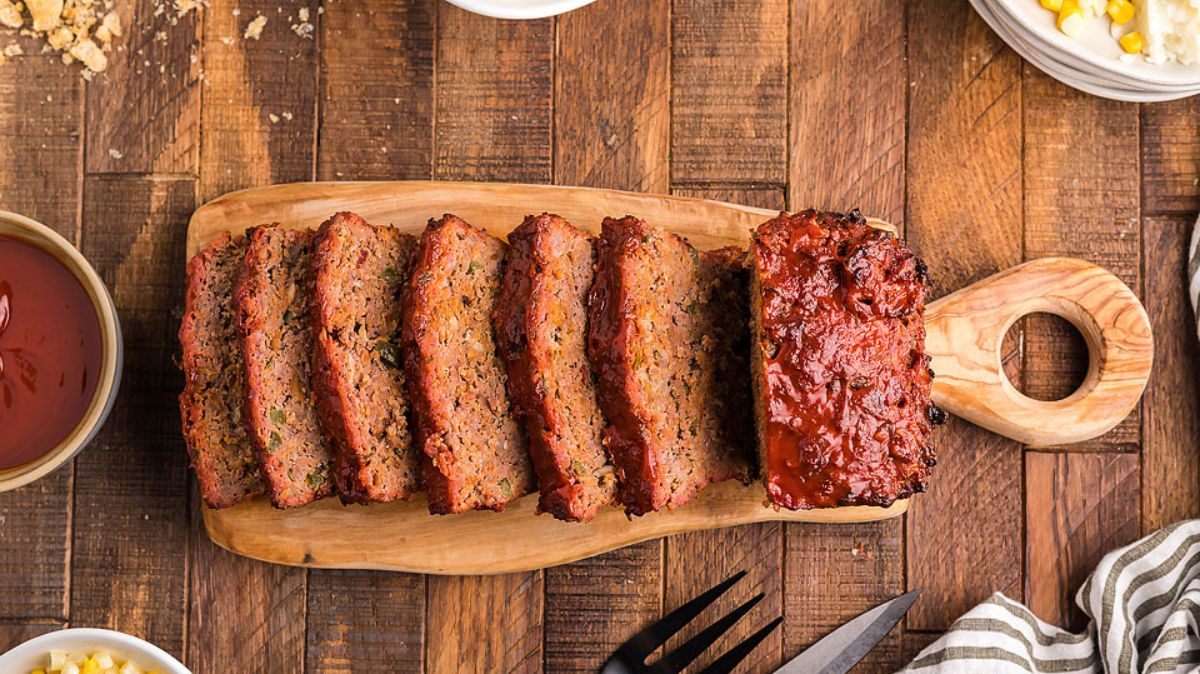 Sliced meatloaf arranged on a wooden cutting board, with a serving fork and bowls of food nearby on a wooden table.