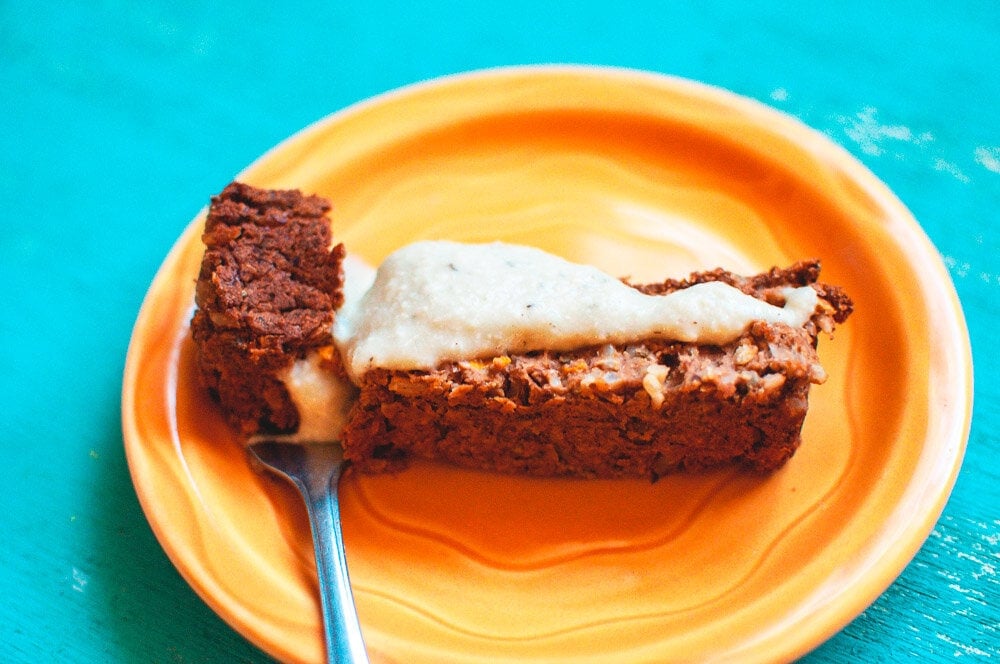 A slice of brown cake with light-colored frosting on an orange plate, with a fork resting on the plate, on a turquoise surface.