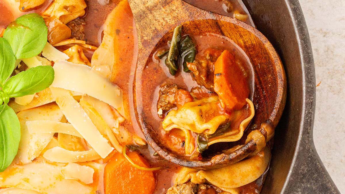 A close-up of a pot of lasagna soup with visible pasta, sliced carrots, ground meat, spinach, and tomato broth, garnished with basil and cheese shavings.