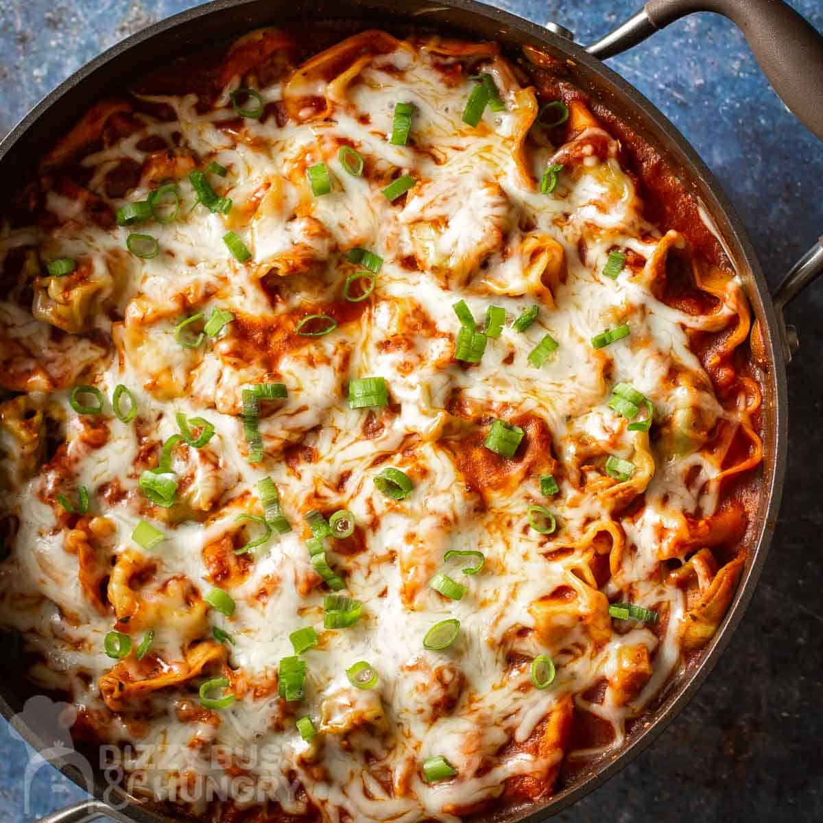 A skillet filled with baked lasagna noodles, tomato sauce, melted cheese, and chopped green onions on top.