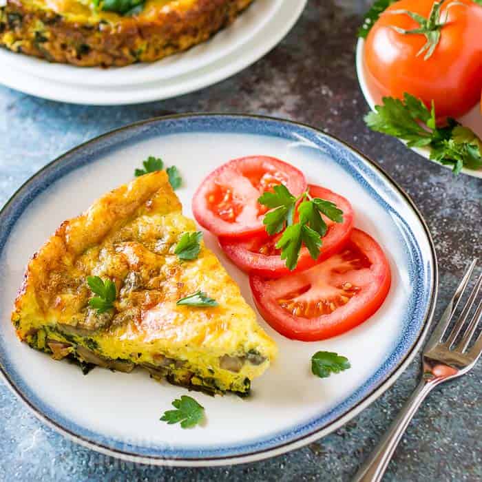 A slice of vegetable frittata garnished with parsley is served with sliced tomatoes on a blue-rimmed plate. A fork and fresh tomatoes are nearby.
