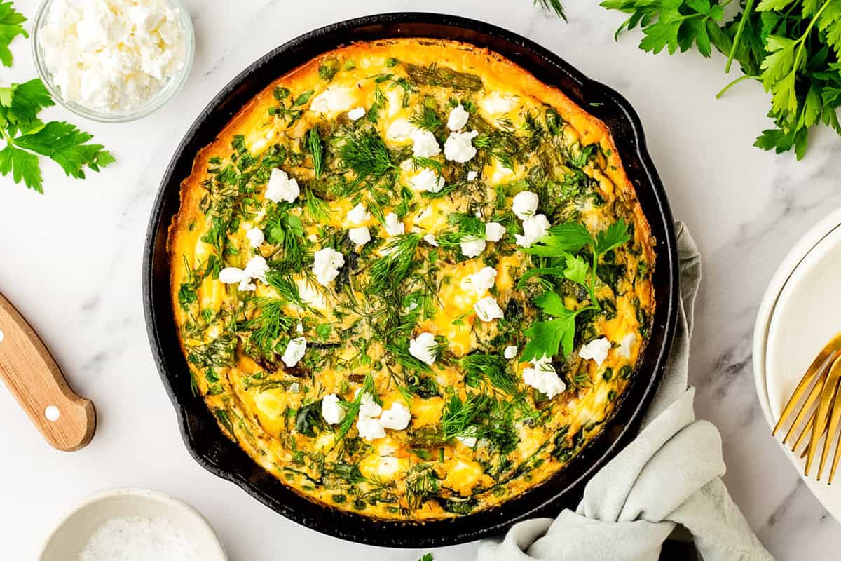 A baked vegetable frittata topped with fresh herbs and crumbled cheese in a cast iron skillet, surrounded by parsley, a dish of cheese, and plates with gold forks.