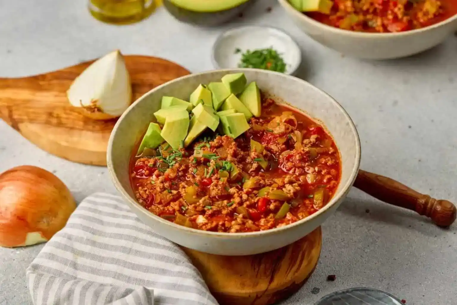 A bowl of chili topped with diced avocado, placed on a wooden board with half an onion, chopped herbs, and a striped cloth nearby.
