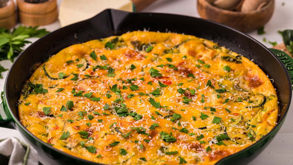 A baked frittata in a black skillet, topped with melted cheese and chopped herbs, sits on a kitchen counter alongside fresh ingredients.