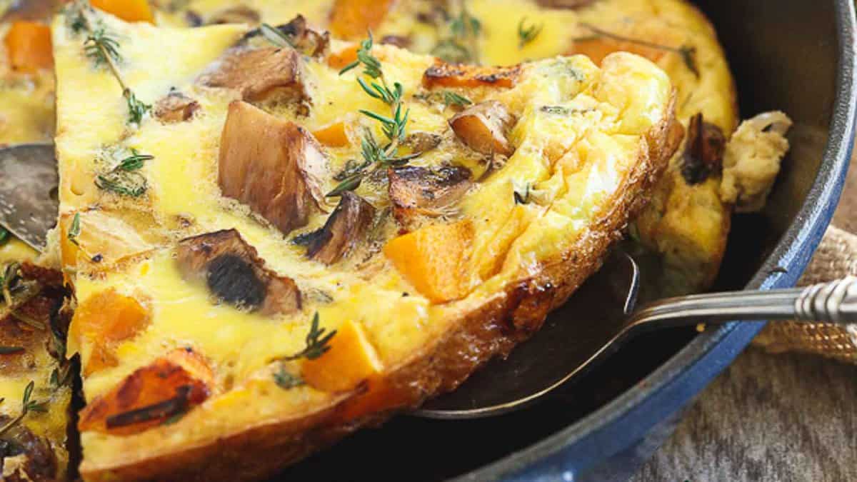 A slice of vegetable frittata with mushrooms and herbs is being served from a skillet.