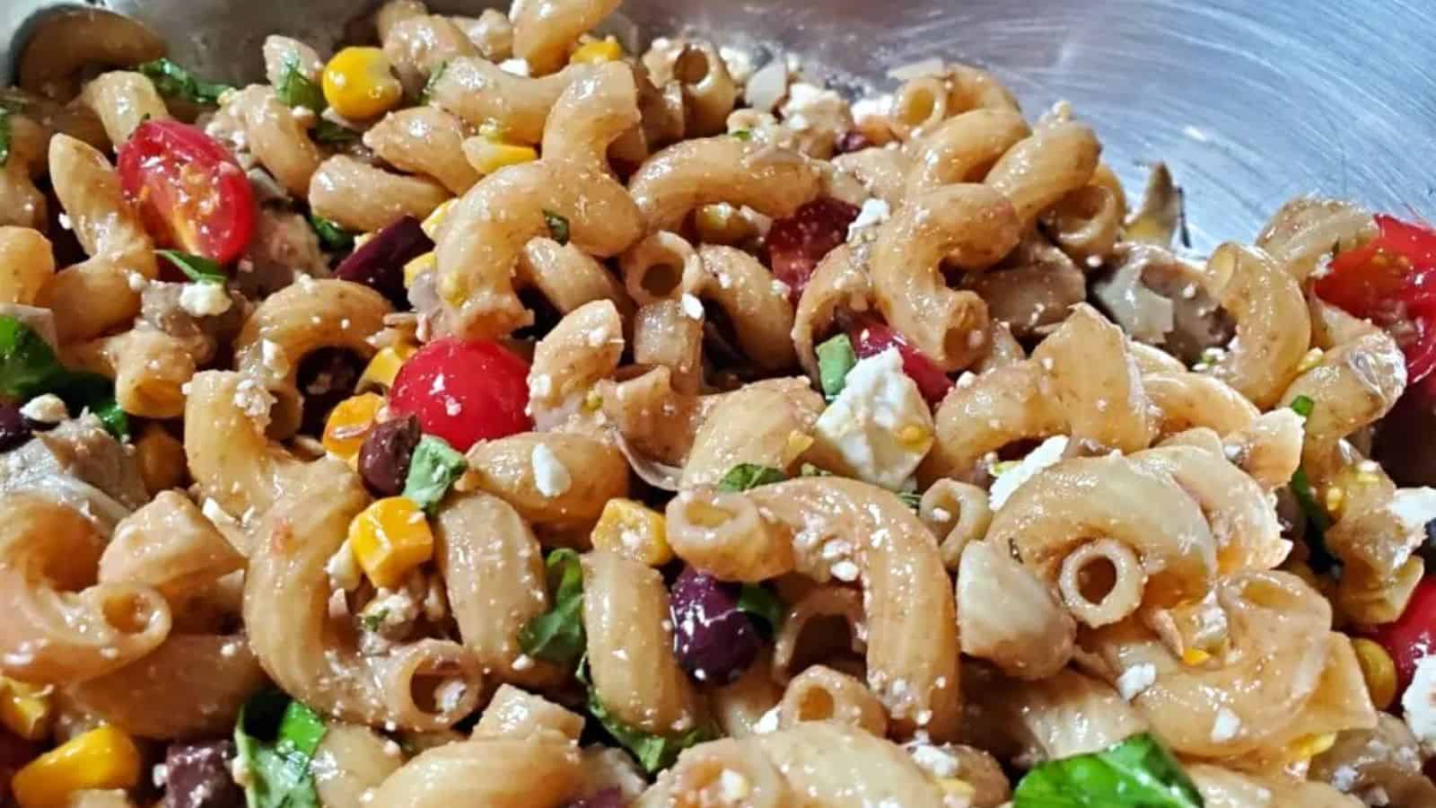 A close-up of pasta salad with elbow macaroni, cherry tomatoes, corn, black olives, spinach, and crumbled feta cheese in a metal bowl.