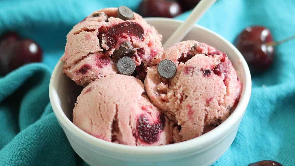 A white bowl with three scoops of cherry ice cream containing dark chocolate chips, placed on a blue cloth with cherries in the background.