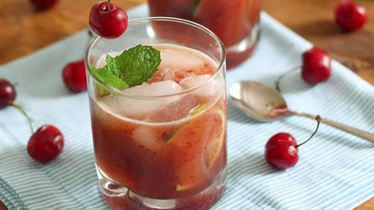 A glass of iced cherry drink garnished with a mint sprig and a cherry, placed on a striped napkin, with cherries and a spoon nearby.