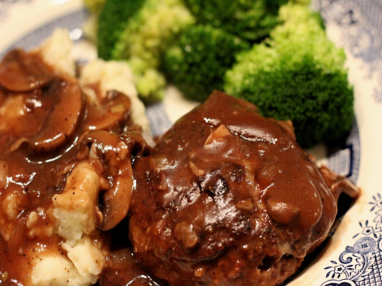 Plate of mashed potatoes with brown mushroom gravy, Salisbury steak topped with brown sauce, and steamed broccoli florets.
