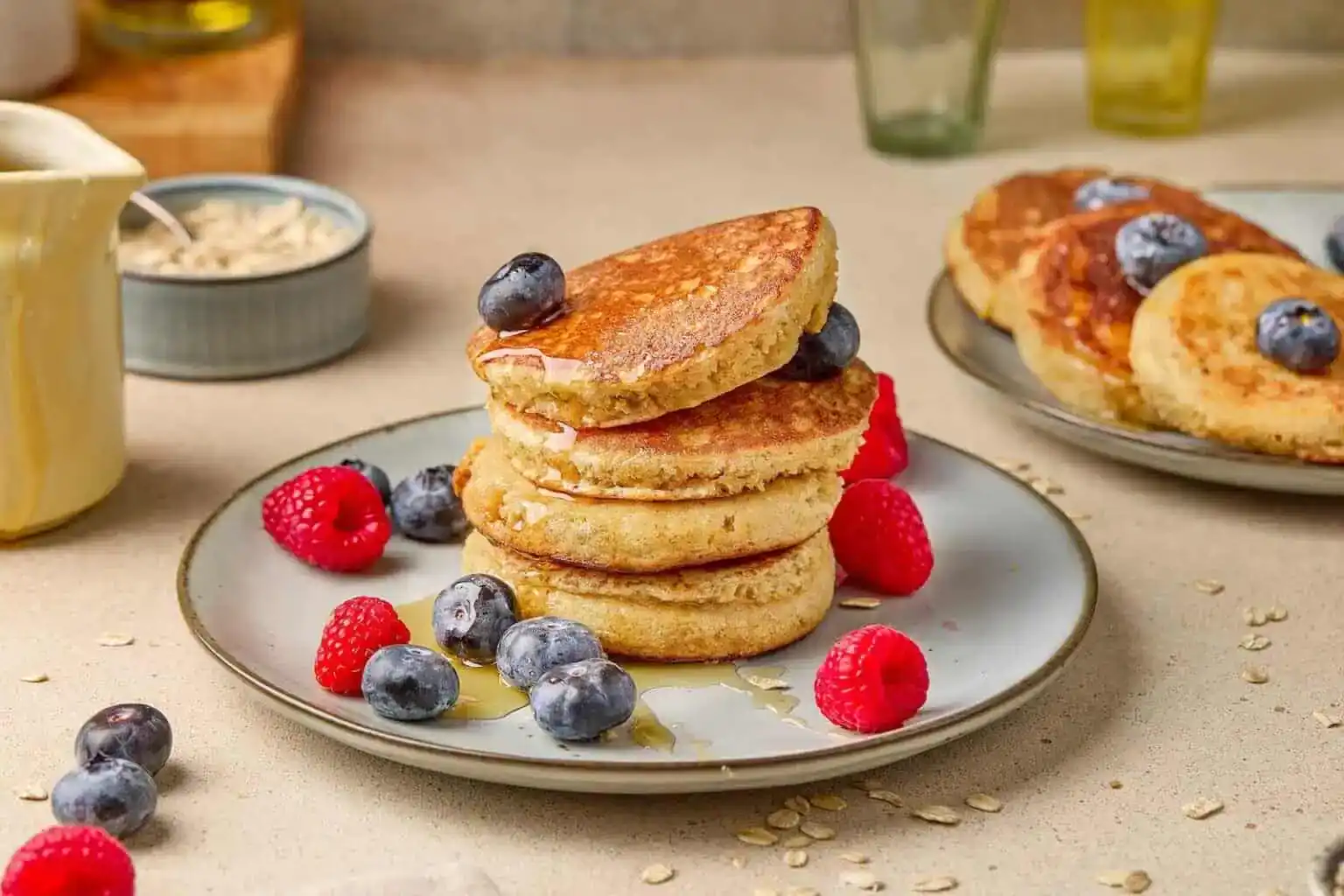 A plate of thick pancakes topped with blueberries, raspberries, and honey, with another plate of pancakes and some glasses in the background.