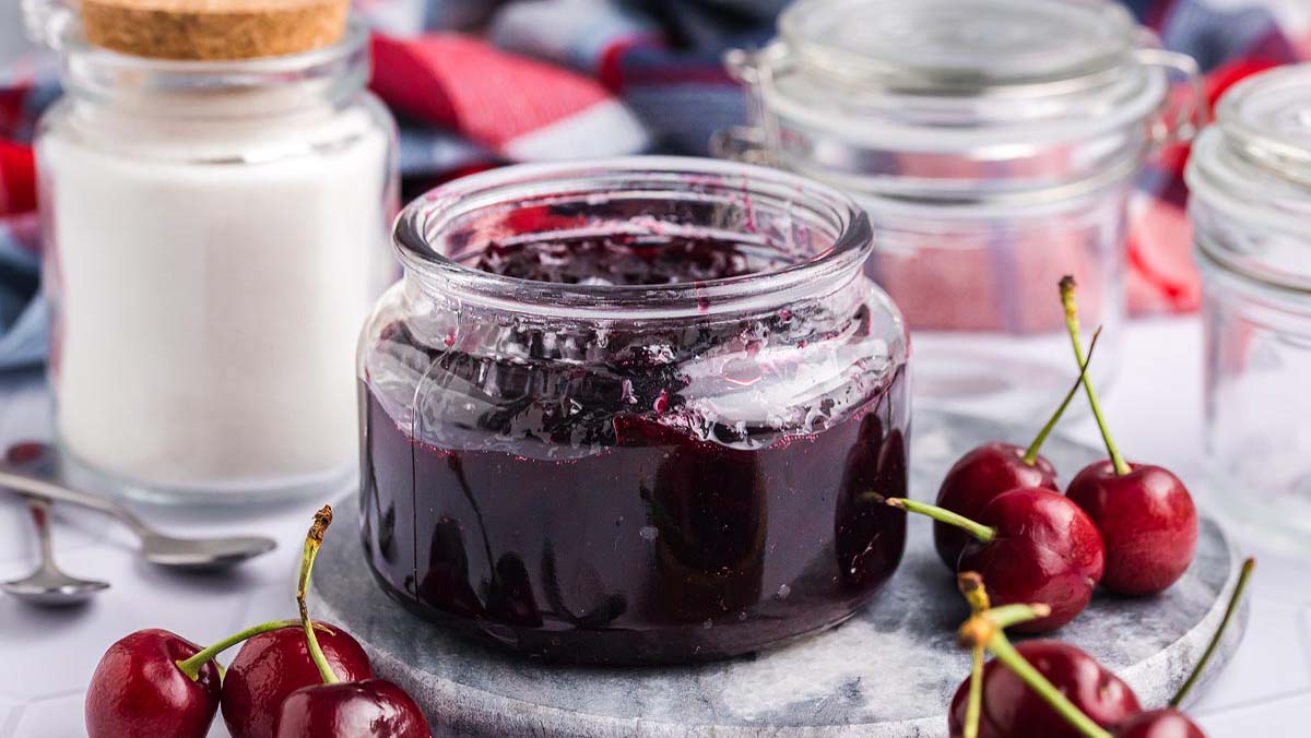 A glass jar filled with cherry jam sits on a stone coaster, surrounded by fresh cherries and glass jars, one containing sugar.
