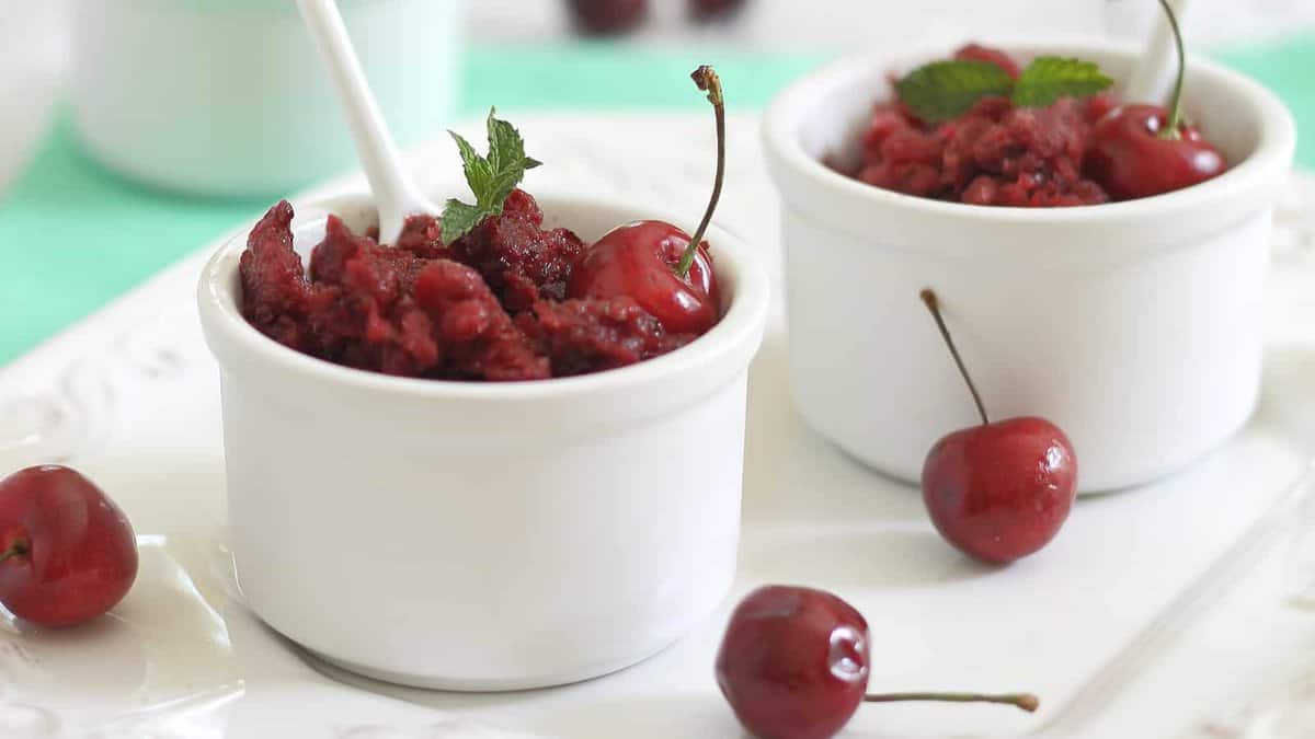 Two white ramekins filled with cherry granita, garnished with mint, are placed on a tray with fresh cherries scattered around.