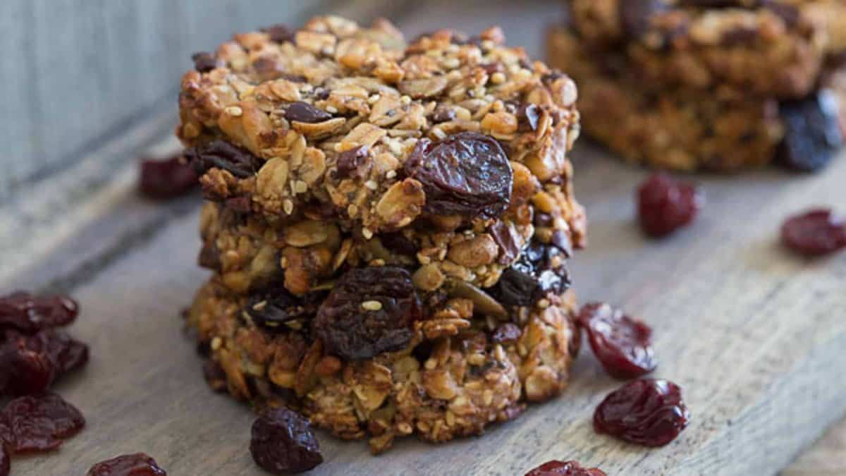A stack of homemade granola cookies with seeds, oats, and dried cranberries, placed on a wooden surface with more cranberries scattered around.