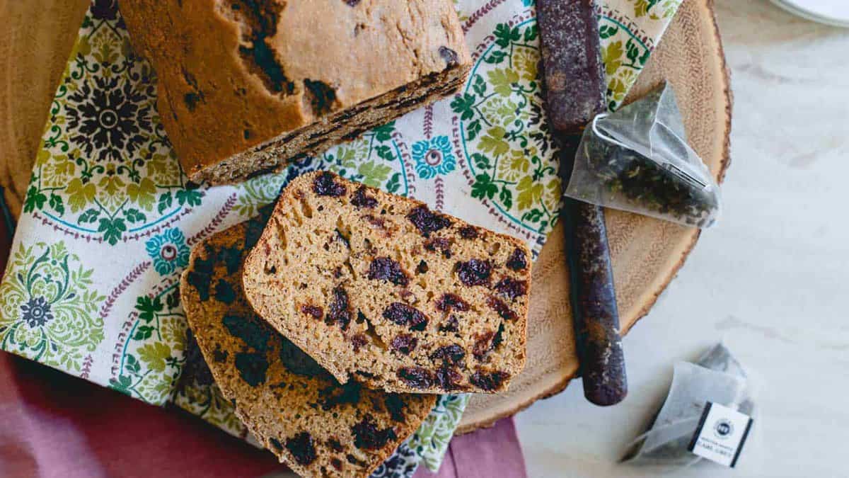 A sliced loaf of fruit-studded bread sits on a patterned cloth with a knife and two tea bags beside it.