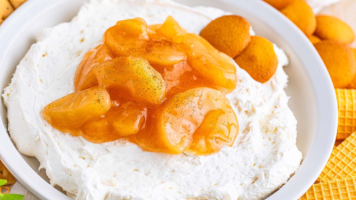 A bowl filled with creamy white dip topped with spiced apple slices, surrounded by vanilla wafer cookies and waffle cookies.